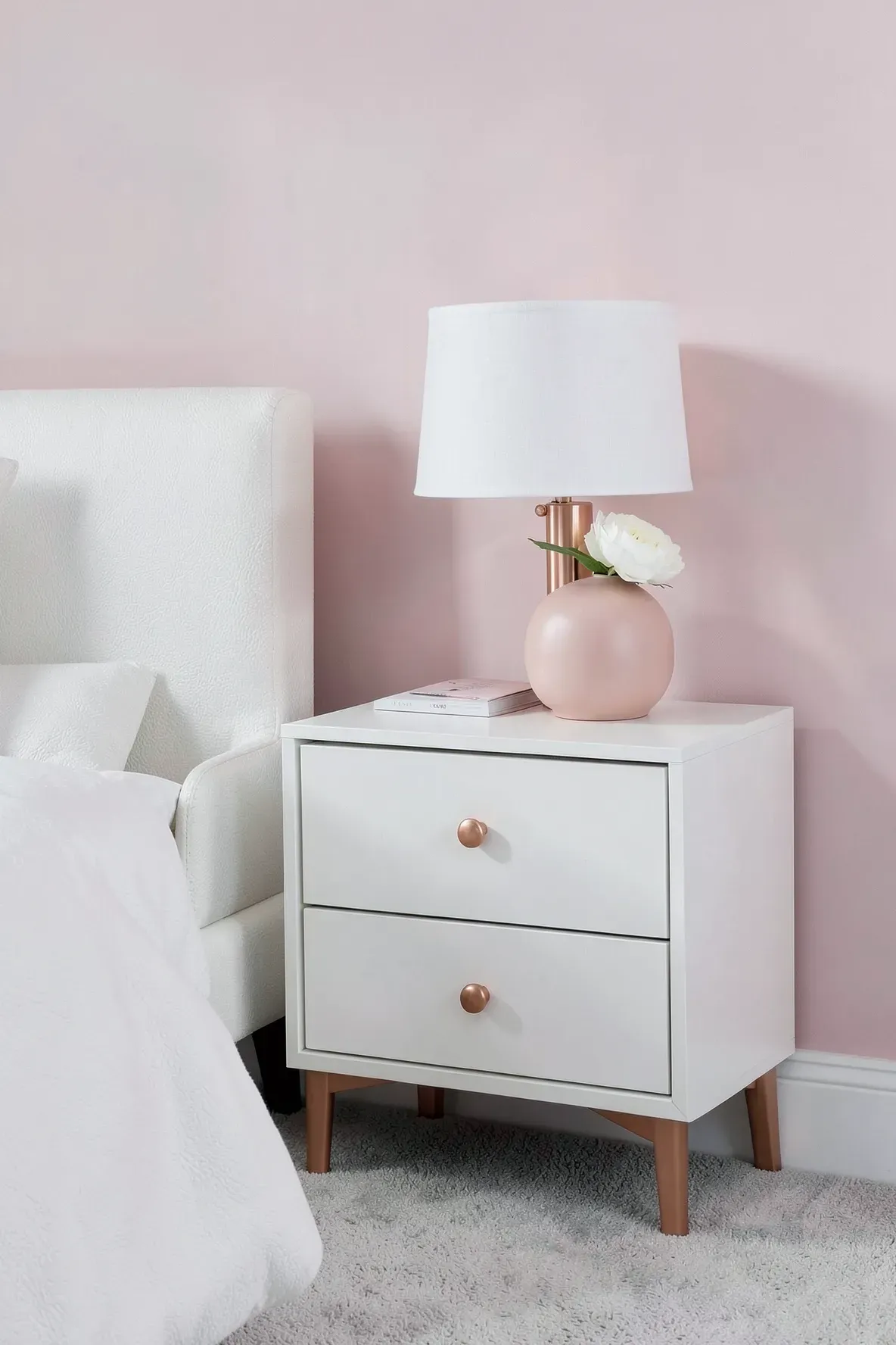 A matte white nightstand with rose gold knobs and a matching lamp against a pink wall.