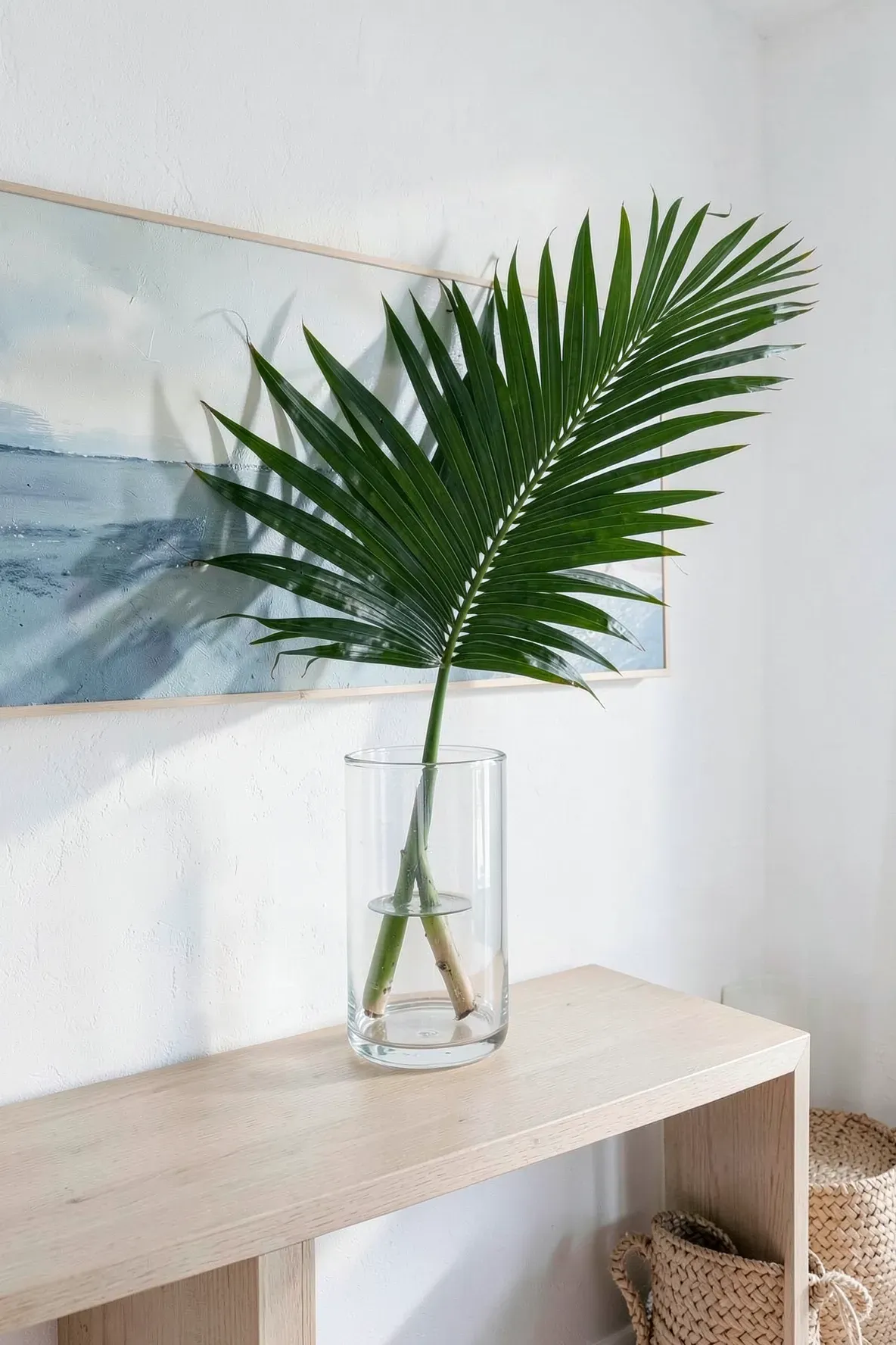 A single large green palm leaf in a clear glass vase on a light wood console table.