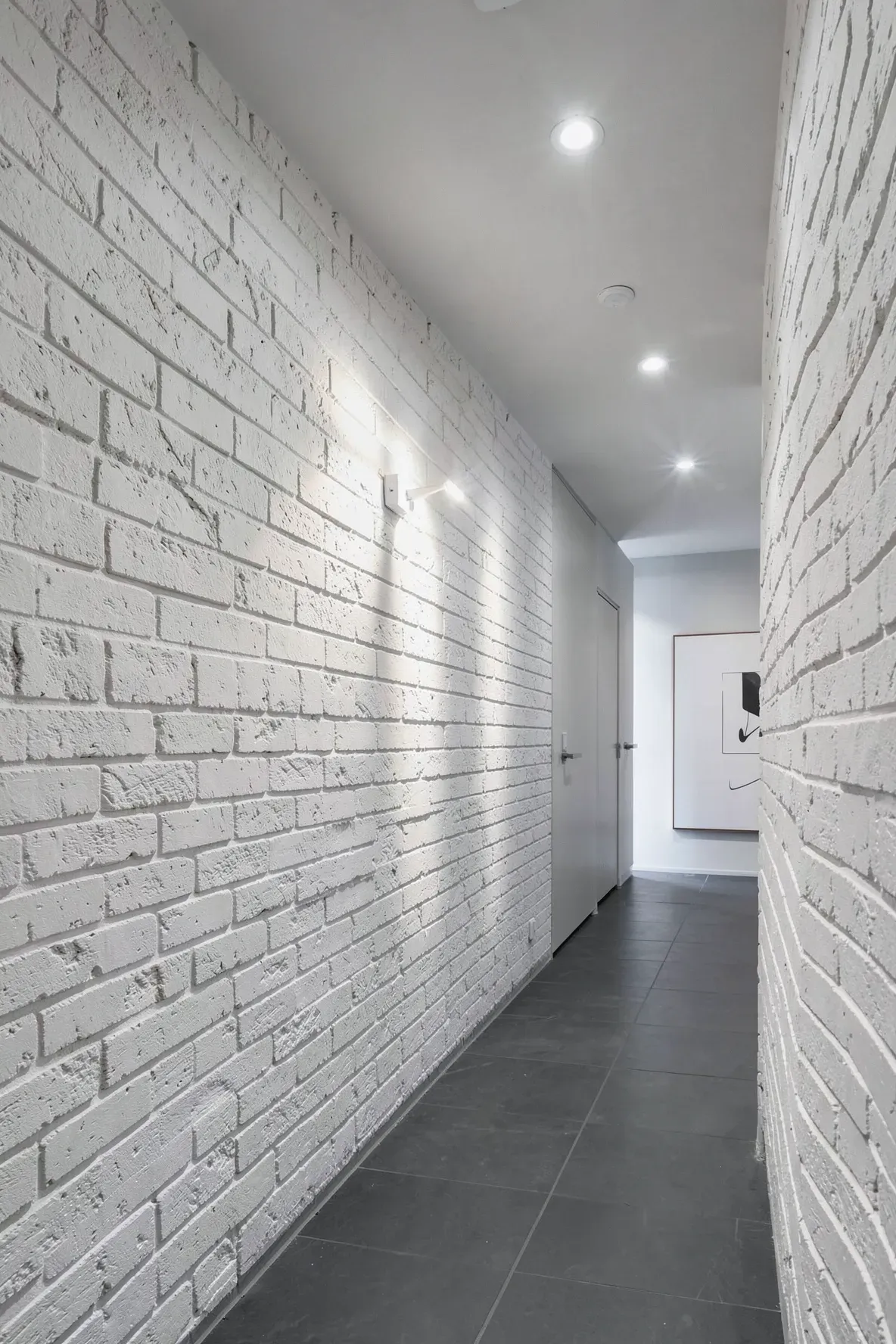 A hallway with ceiling lights washing a white brick wall to highlight its texture and depth.