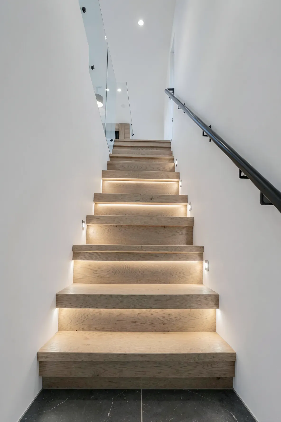 Recessed square LED lights illuminating the treads of a modern wooden staircase in a hallway.