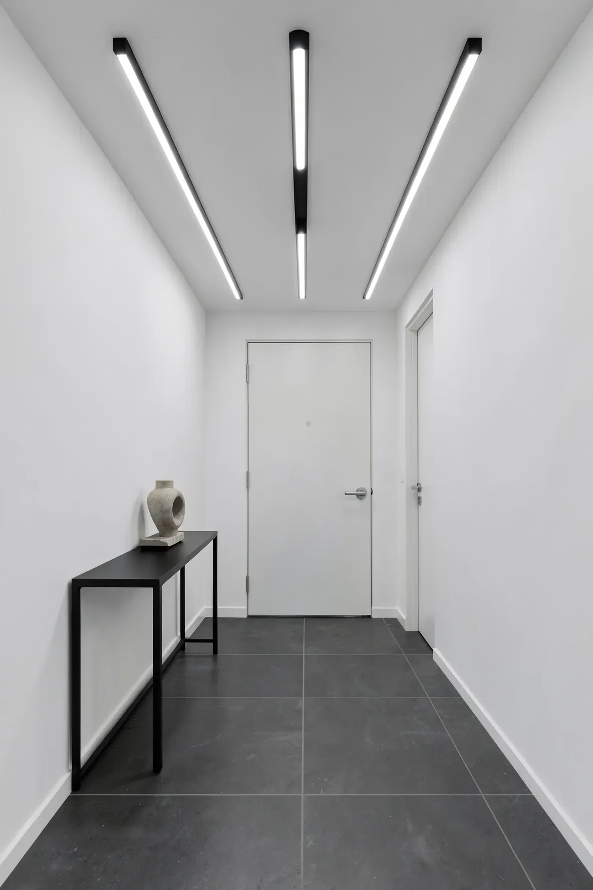 Thin black linear LED light bars on the ceiling of a minimalist white and grey hallway.