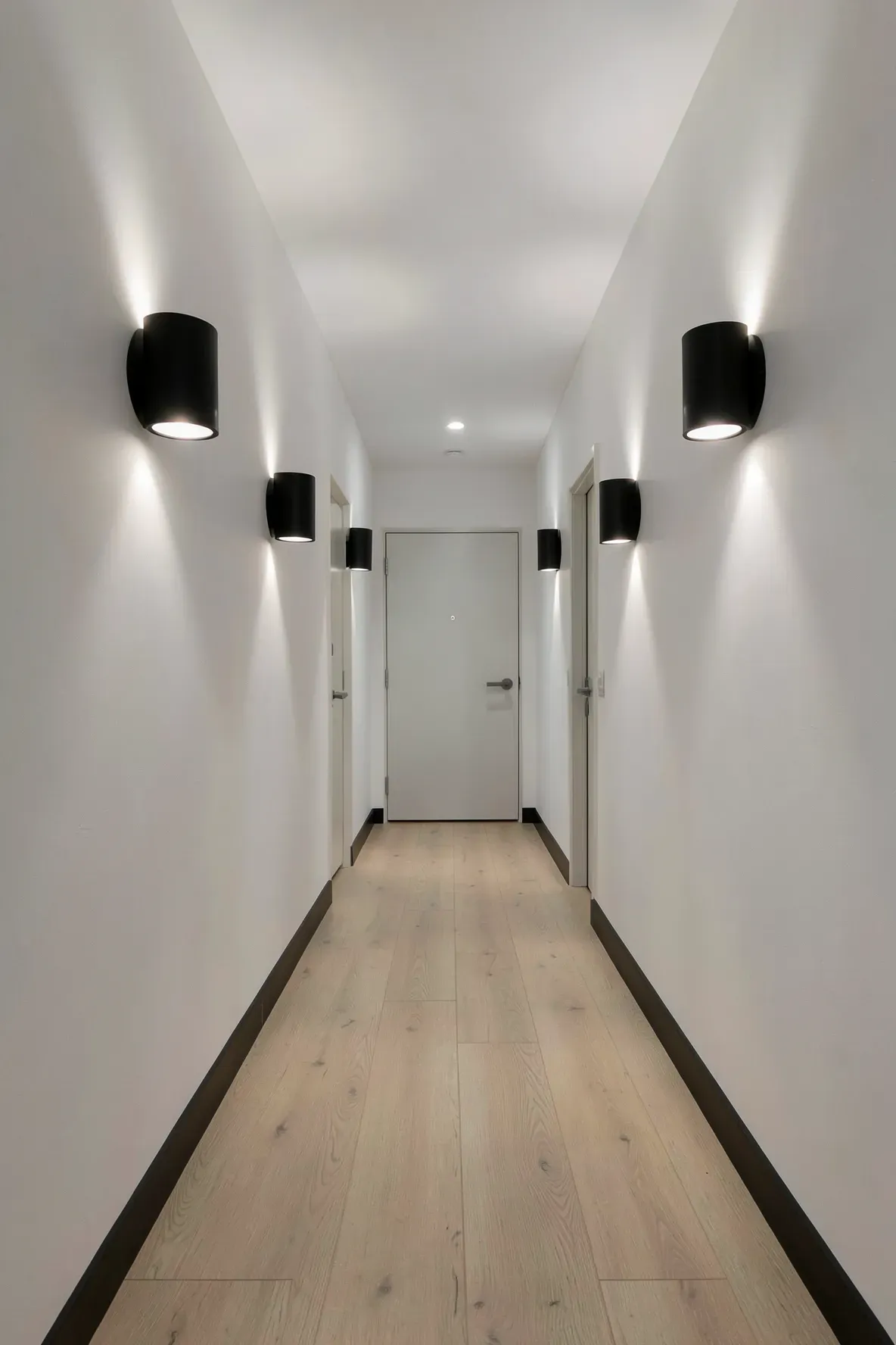 A long narrow hallway with matte black wall sconces casting upward light beams on white walls.