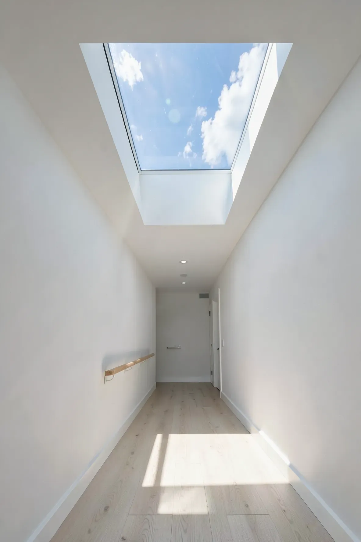 A bright hallway with a large ceiling skylight letting in direct sunlight onto light wood floors.