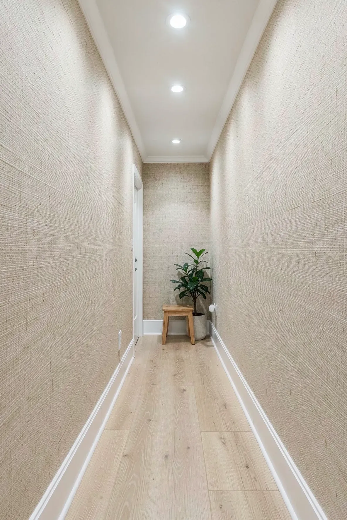A narrow hallway with beige textured grasscloth wallpaper and light wood floors.