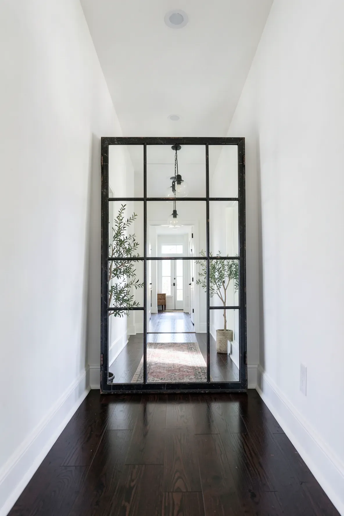 A large black window pane mirror at the end of a white hallway reflecting the entire space.
