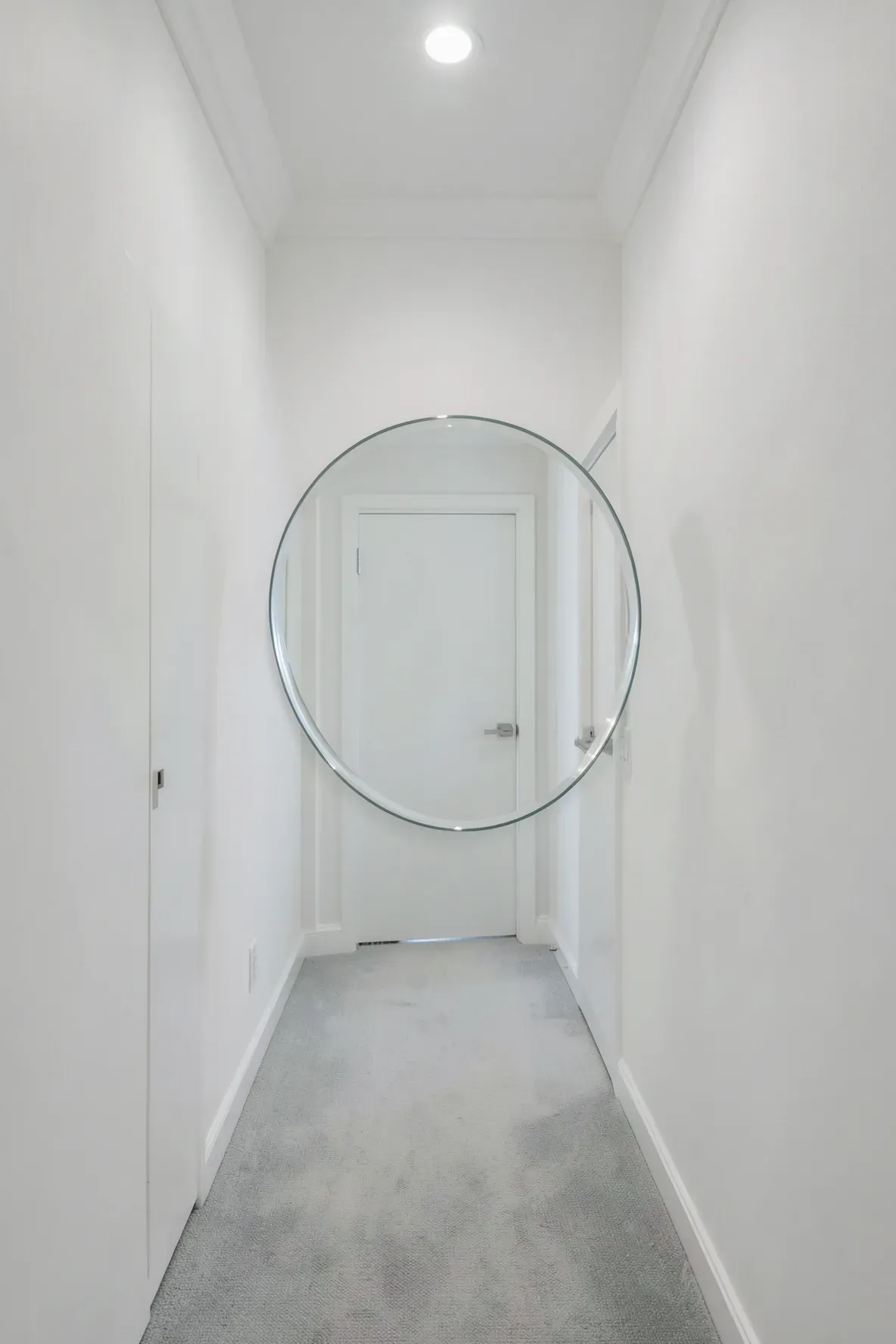 A frameless round mirror with a beveled edge hangs in a clean, minimalist white hallway.