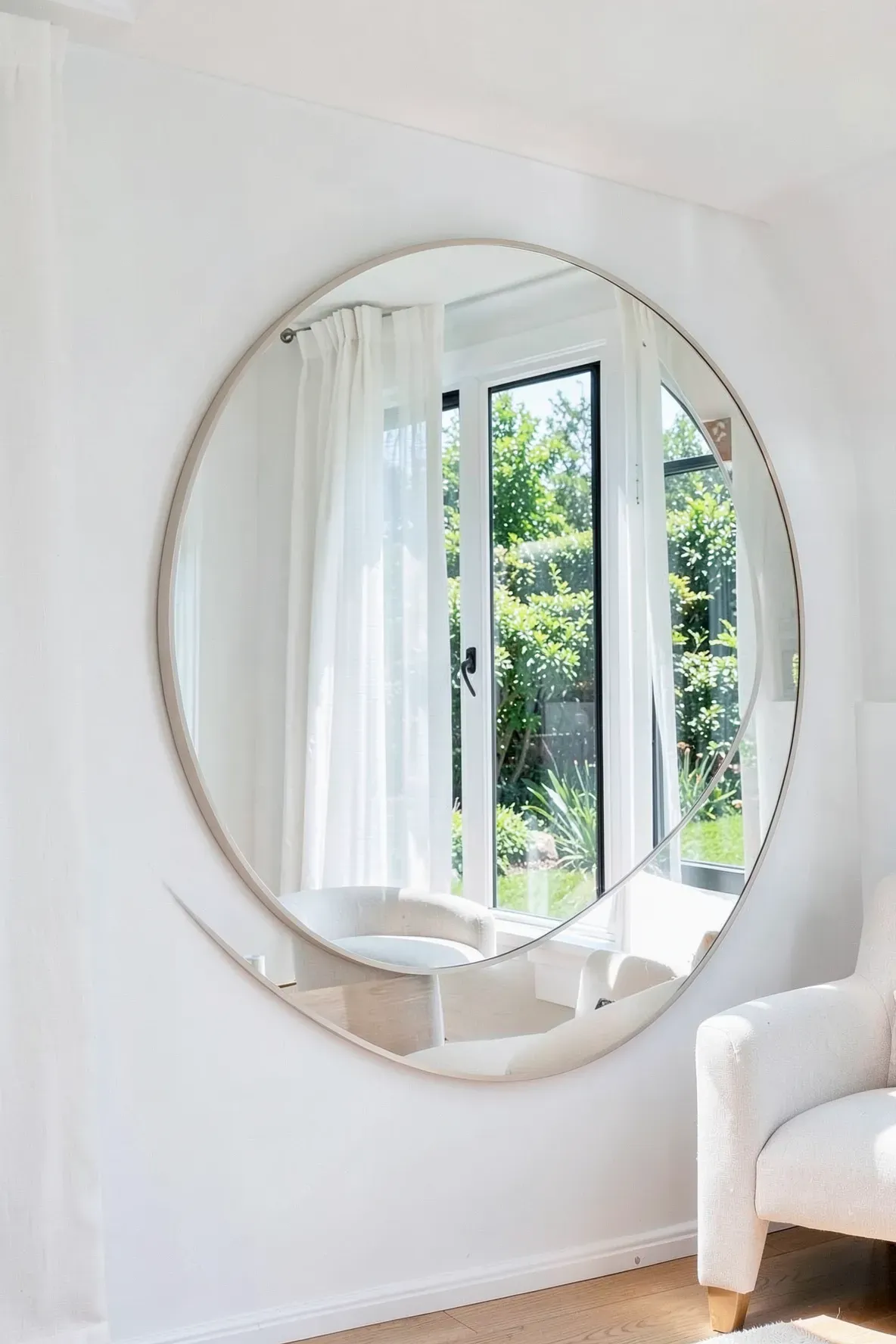 A large round mirror hangs on a white wall, reflecting a bright window and a green garden.