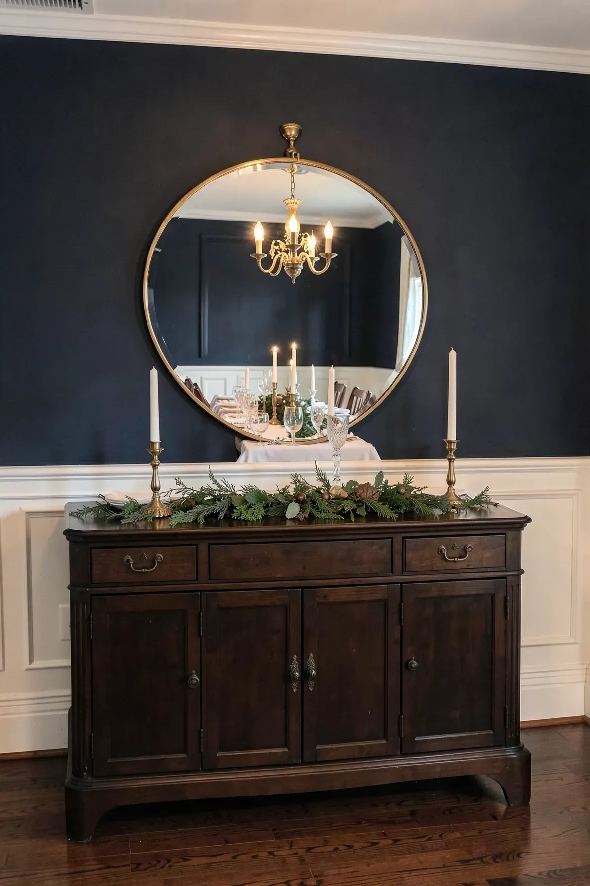 A large round mirror hangs above a dark wood buffet in an elegant dining room with navy walls.