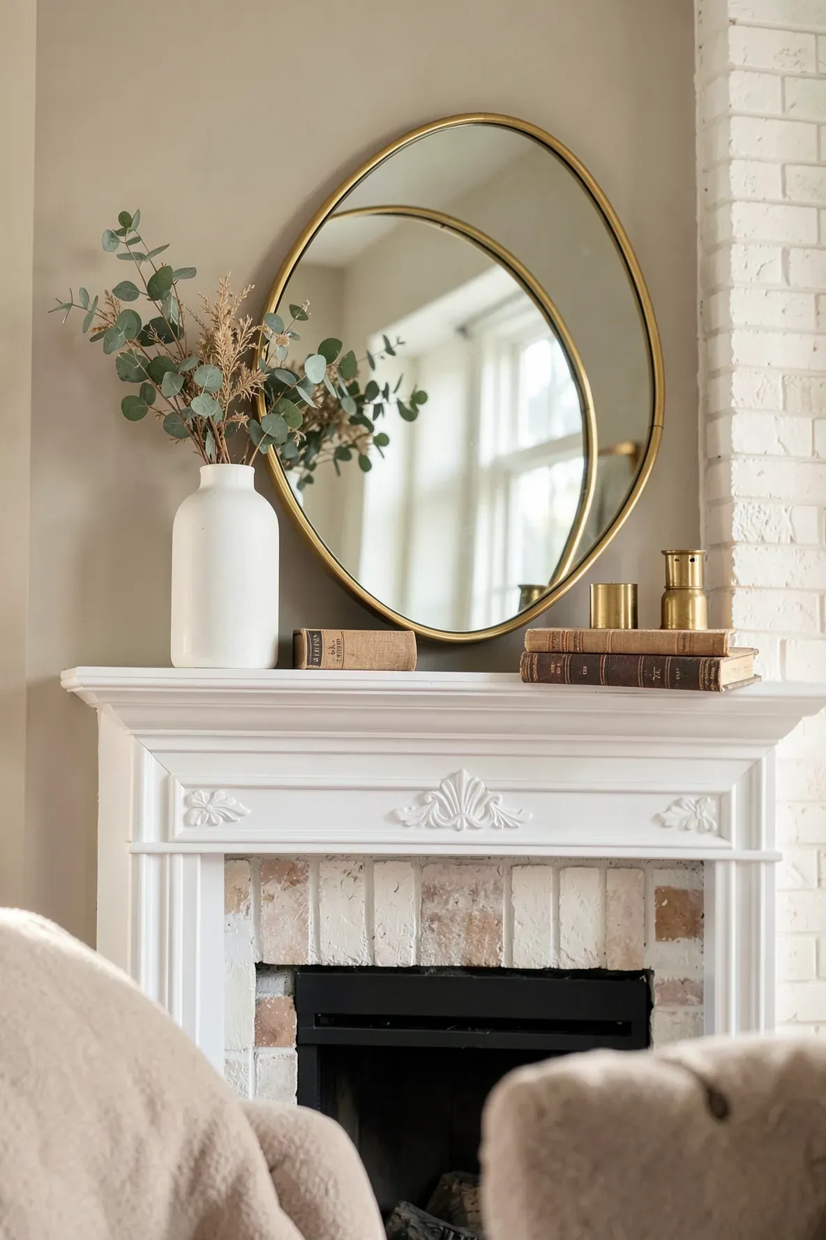 A round gold mirror leans on a white fireplace mantle with a vase and books.