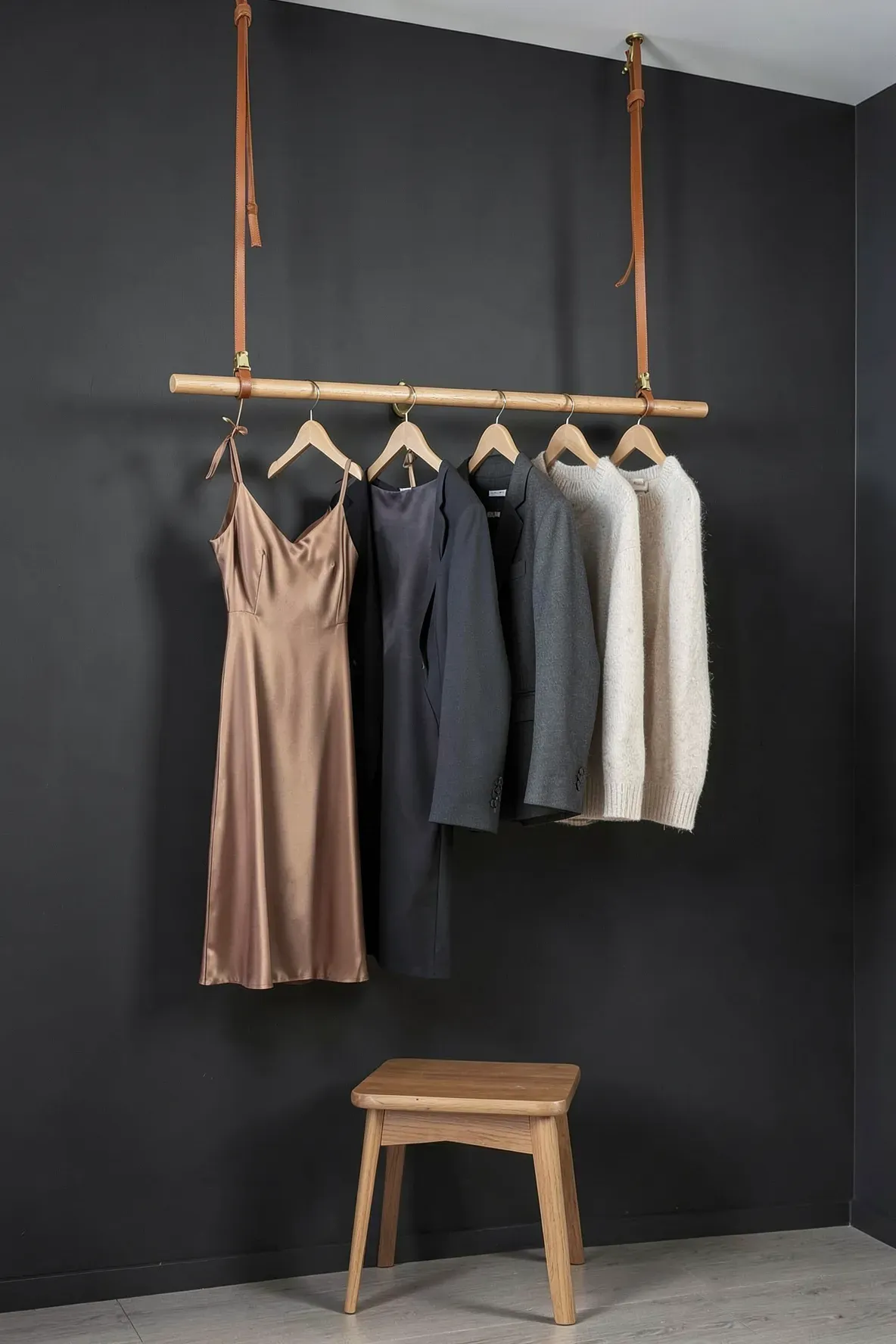 A minimalist wooden dowel hanging from leather straps on a dark wall with boutique clothing.