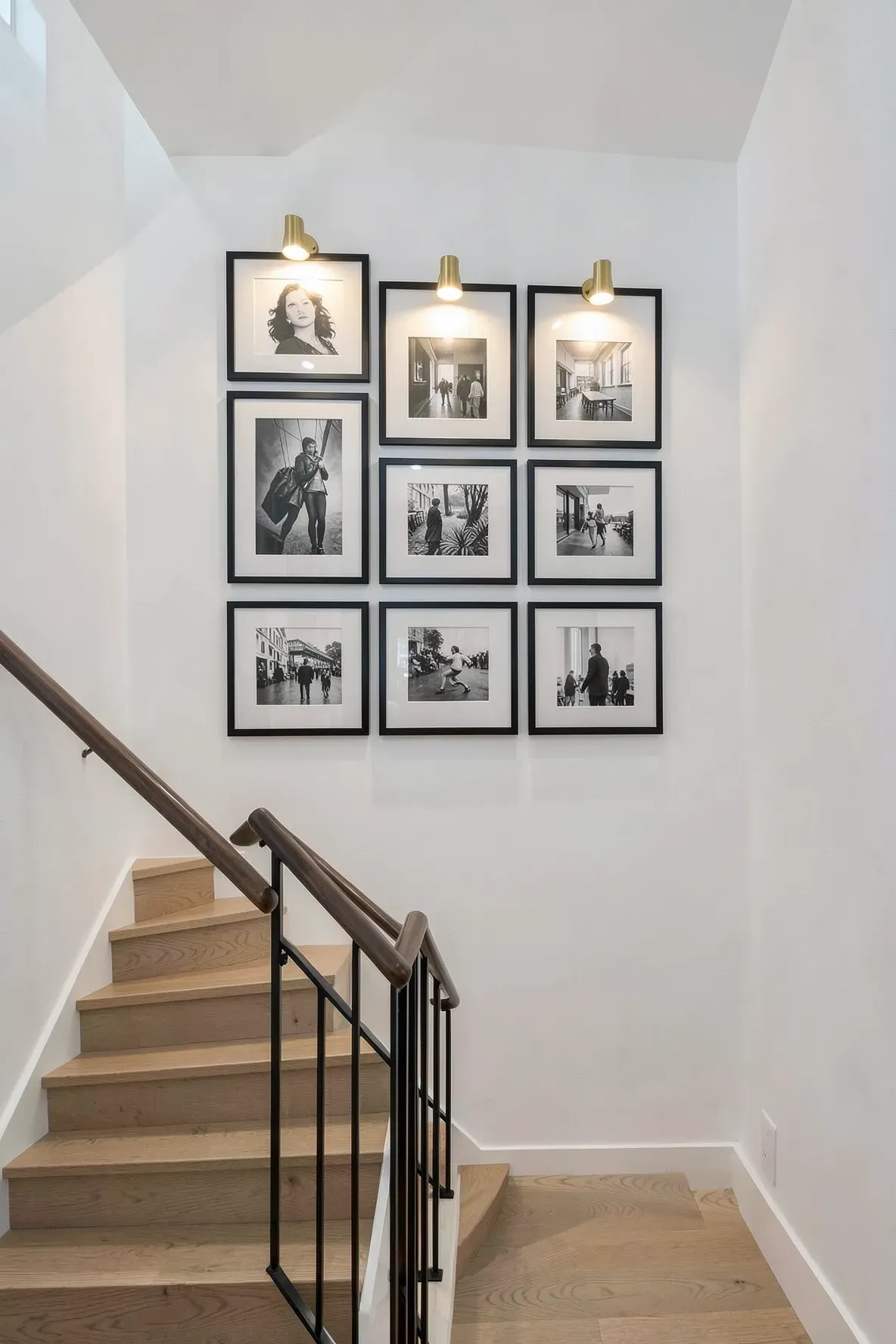 A symmetrical 3x3 grid of black frames on a white staircase wall with brass picture lights.
