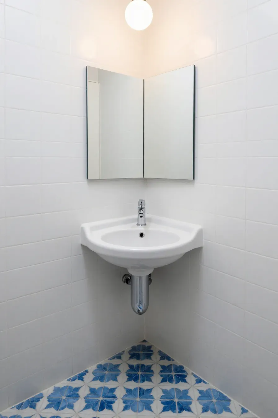 A compact white corner sink and mirrored cabinet in a small bathroom with patterned floor tiles.