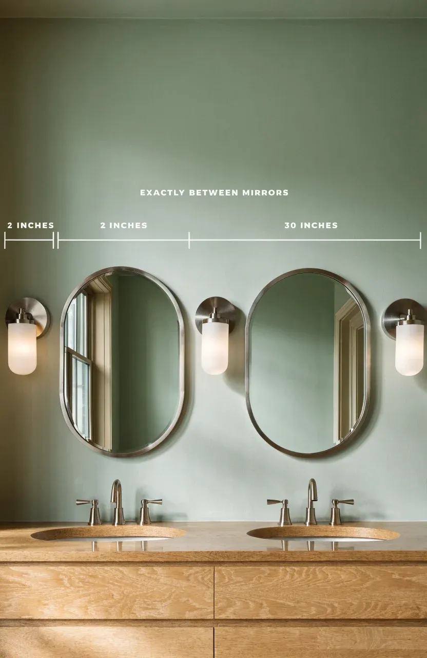 A double vanity with three silver sconces spaced evenly around two oval mirrors on a green wall.