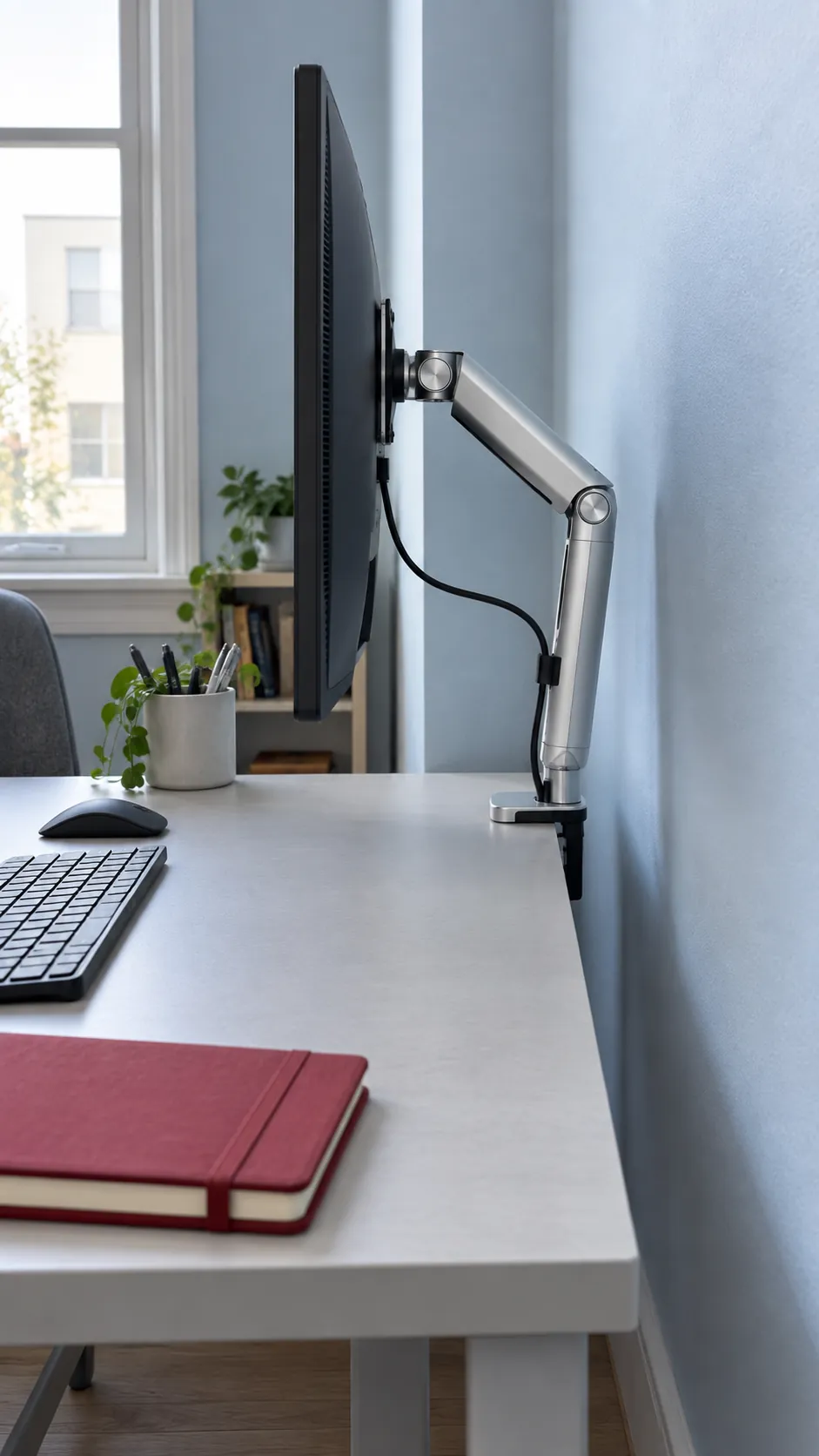 Create a photographic explanation of rear clearance without diagrams: a folded monitor arm seen from the side of a shallow desk, with visible air between the elbow and the room wall