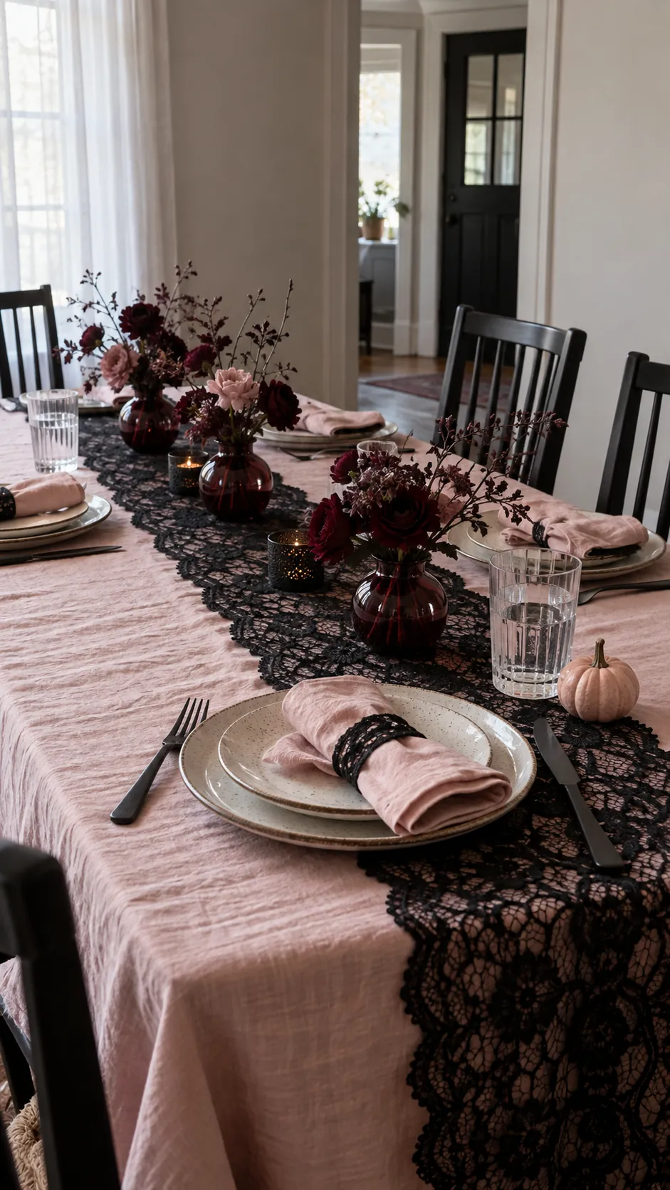 Pink and black Halloween decor with black lace runner laid across the cloth, simple ceramic place settings, and low burgundy bud vases