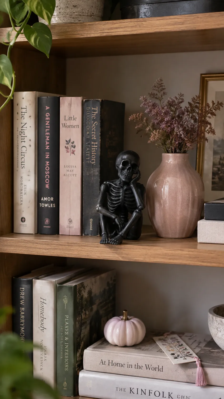 Pink and black Halloween decor with one small black skeleton figurine sitting beside books and a blush ceramic vase