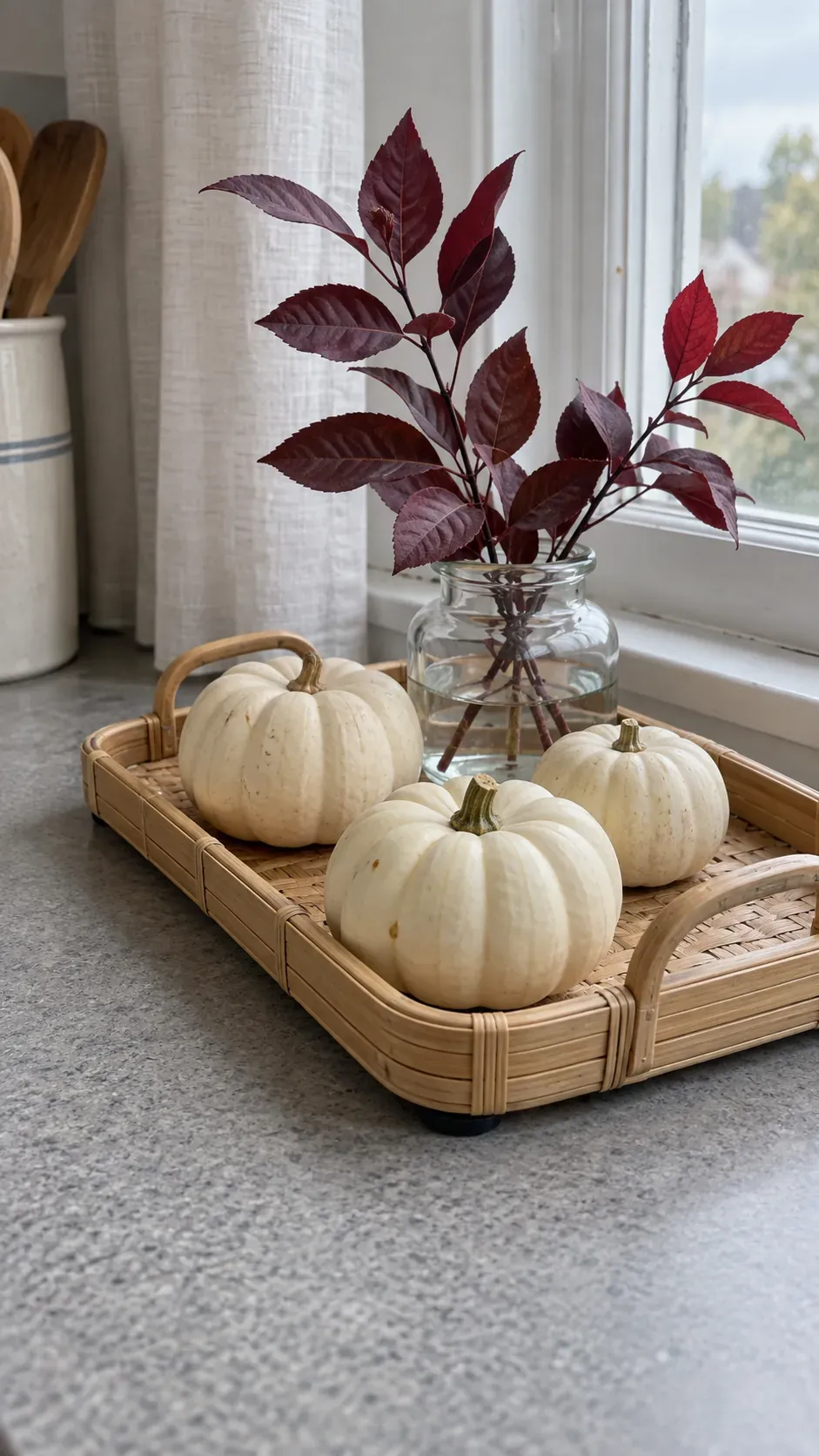Three small ivory pumpkins and a squat glass foliage jar on a shallow bamboo tray beside an open food preparation surface