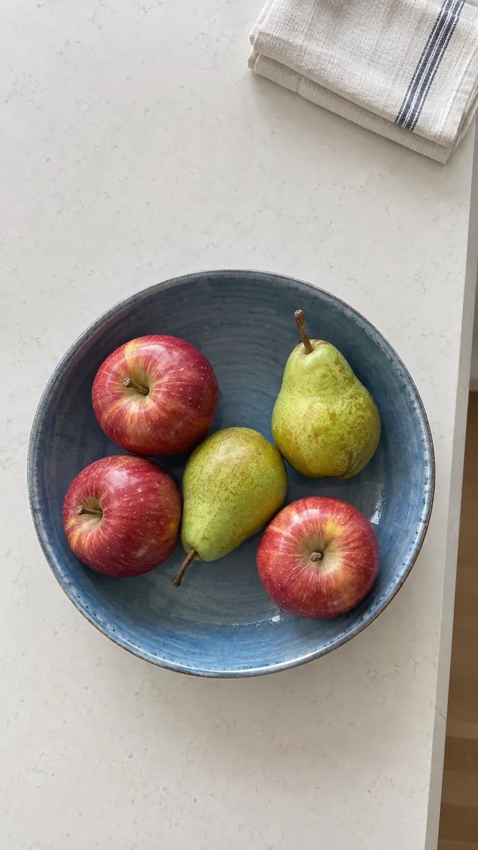 A shallow blue ceramic bowl containing a few red apples and green pears on a breakfast counter