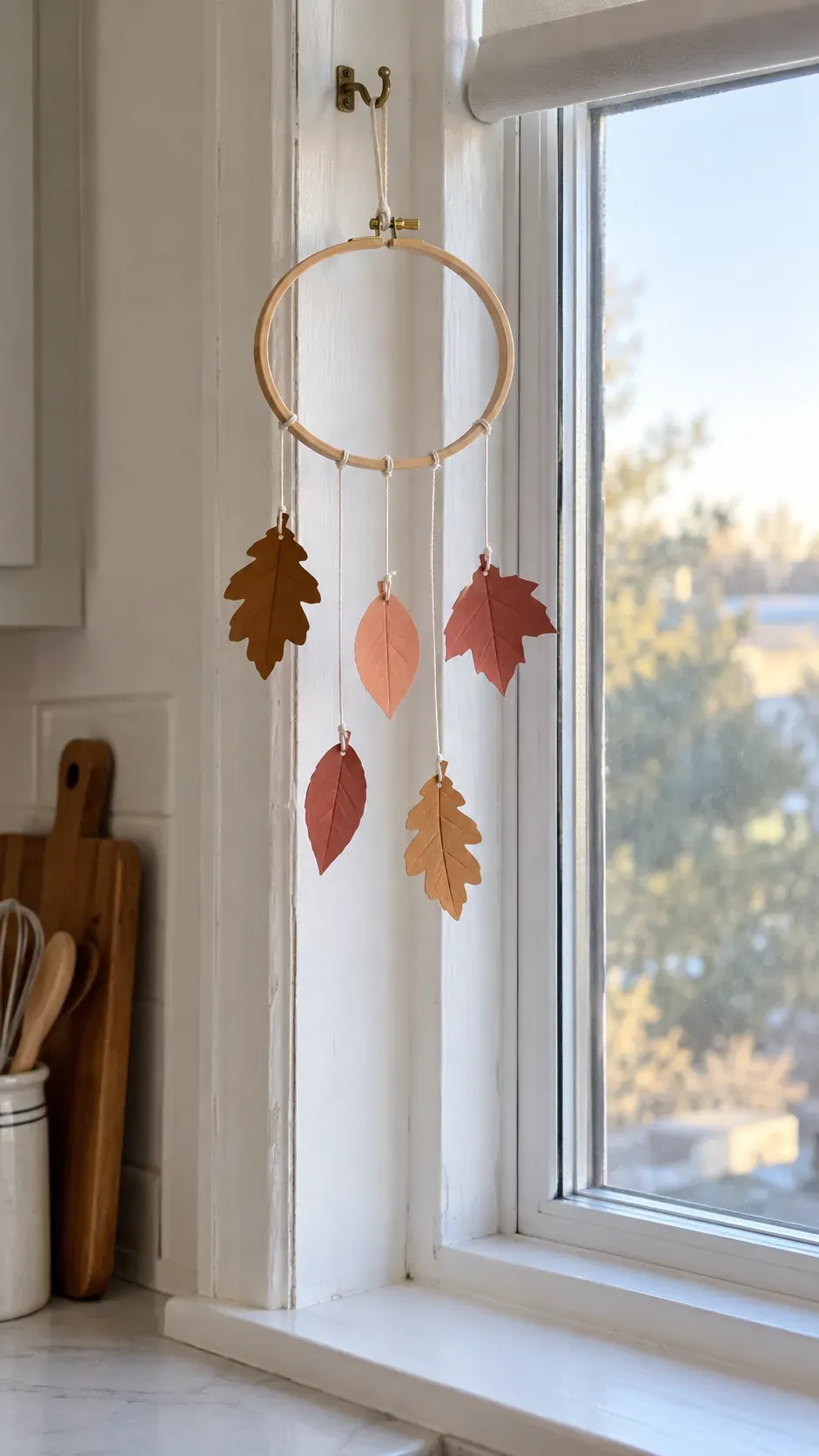 A light wooden hoop mobile carrying paper leaves beside a kitchen window with a clear sill