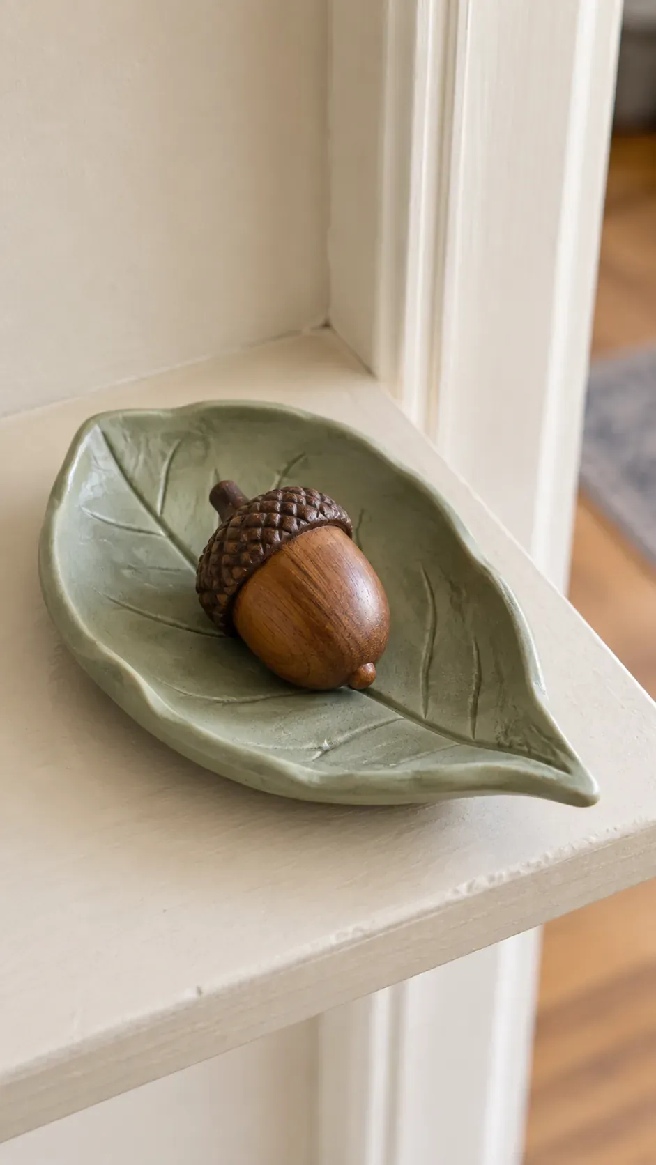 A handmade sage colored clay leaf dish holding one wooden acorn on a kitchen entry shelf
