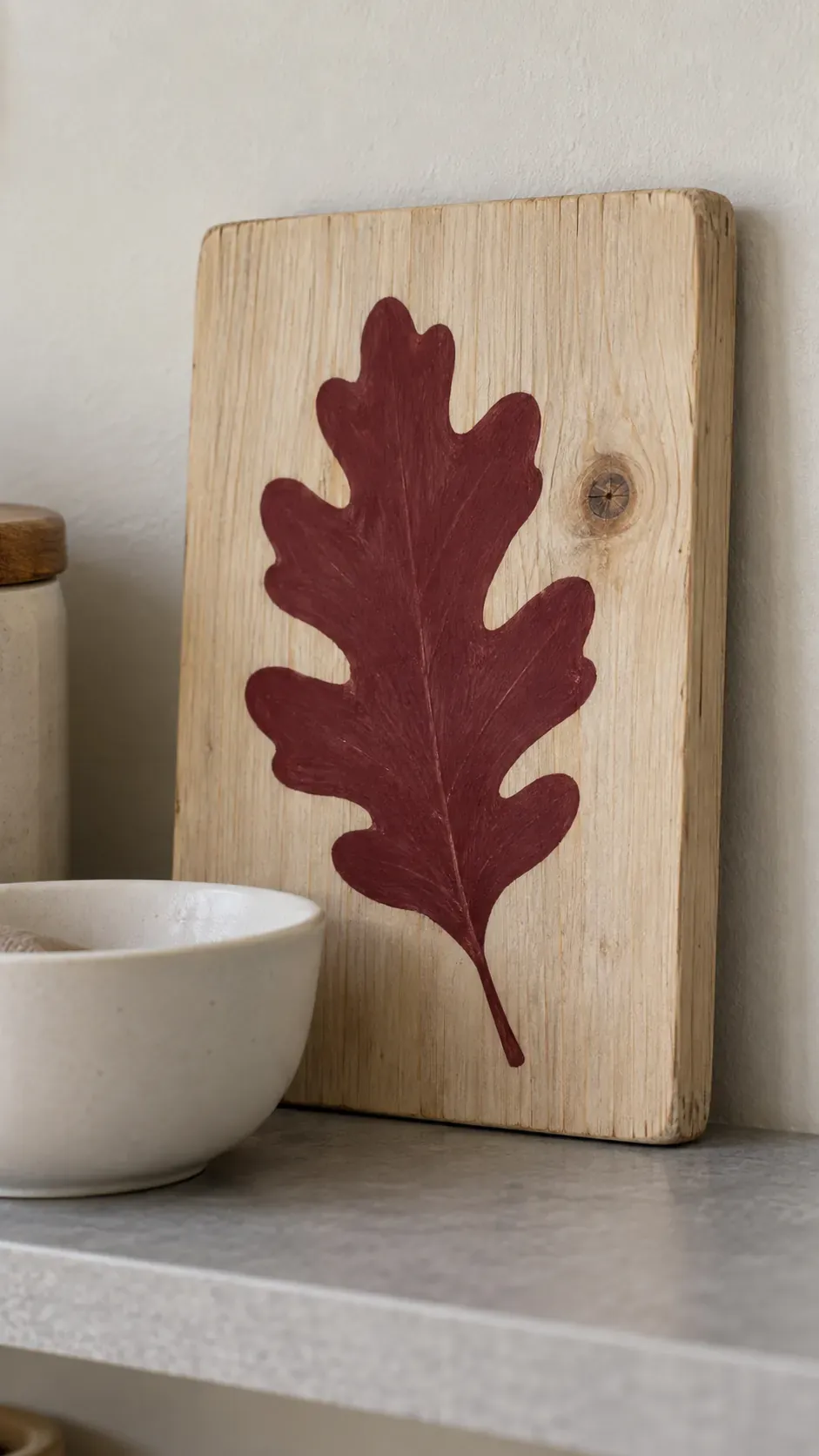 A small salvaged board painted with one burgundy oak leaf silhouette leaning at the back of a shelf