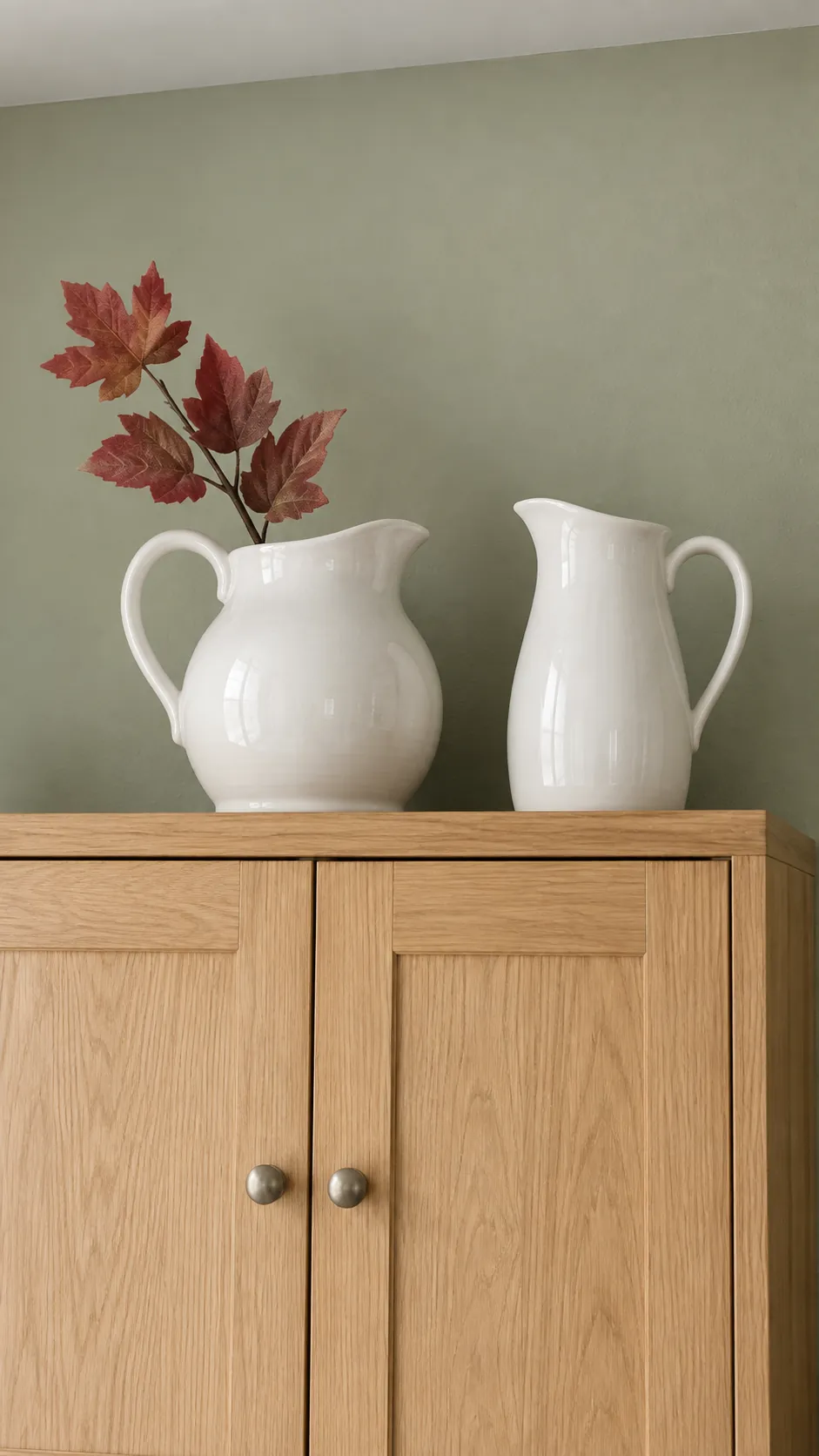Two white pitchers above a cabinet with one short muted red artificial leaf stem