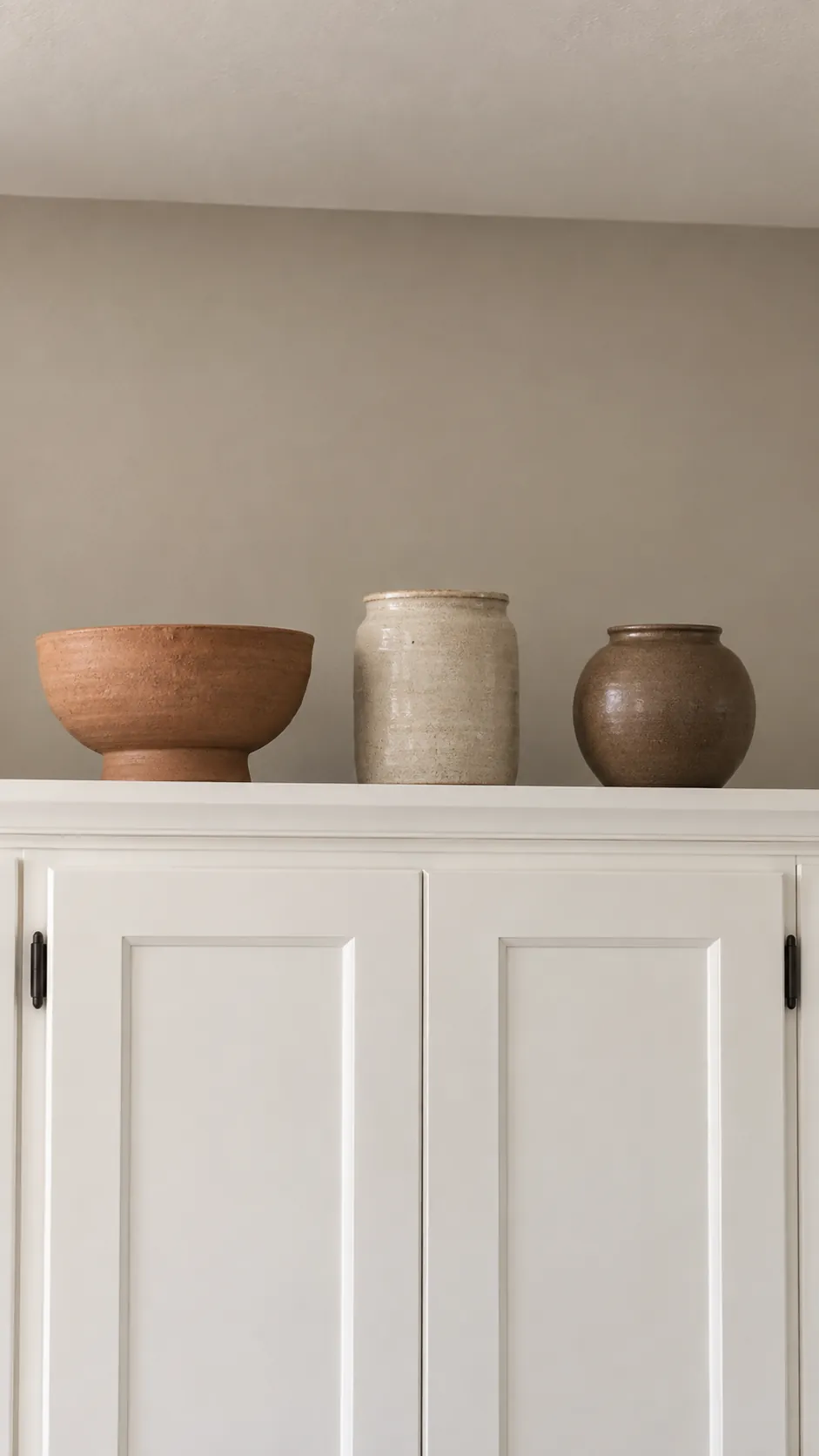 Three unfilled small pottery vessels in clay and oatmeal colors spaced above a cabinet