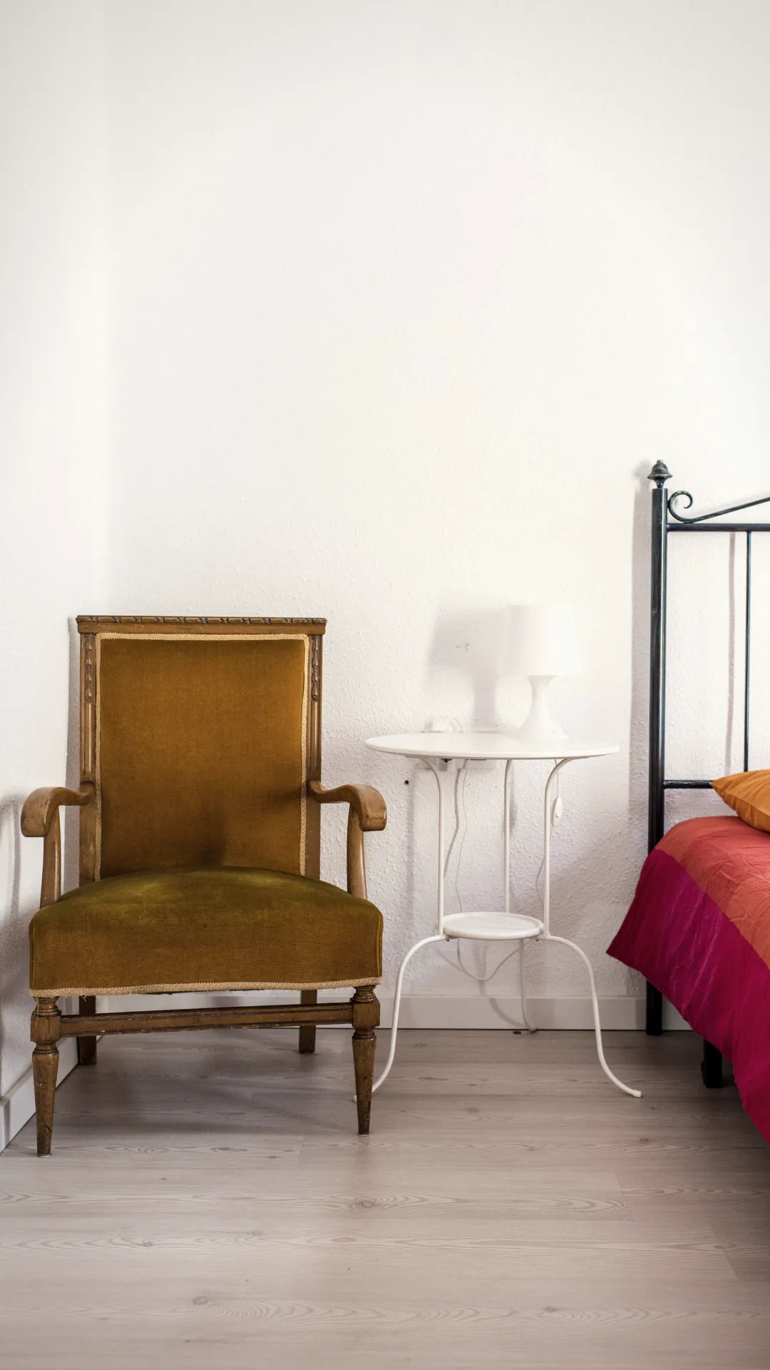 Bedroom reading corner beside the bed with a low armchair, nightstand lamp and stacked books in soft evening light